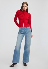 Czerwony dzianinowy top z wysokim kołnierzem i detalami w postaci guzików, połączony z jasnoniebieskimi spodniami z szerokimi nogawkami i czarnymi szpilkami. Gładka faktura, dopasowany krój.