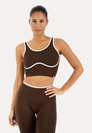 Mujer con top corto ajustado marrón con ribete blanco y leggings de talle alto a juego, de pie con una mano en la cadera contra un fondo liso.