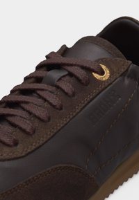 Baskets en cuir marron foncé avec détails en daim, lacets marron passés dans des œillets dorés, et coutures visibles sur le côté.