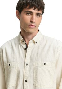 Hellbeiges Hemd mit Knopfleiste aus strukturiertem Stoff, mit zwei Brusttaschen und dunklen Knopfdetails. Kurzer Kragen und dezentes gesprenkeltes Muster.