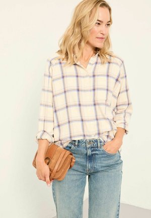 Woman in light plaid shirt and blue jeans holding a small tan leather purse, looking to the side with one hand in pocket.