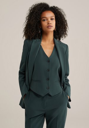 Costum elegant de un verde închis, cu un sacou ajustat, rever în fason clasic și închidere cu nasturi. Sub acesta, o vestă asortată cu decolteu în V și detalii cu nasturi.