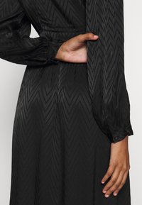 Musta mekko, jossa on chevron-kuvio, pitkät hihat nappikierteillä ja vyötärönauha. Liukas tekstuuri ja vartalonmyötäinen siluetti.