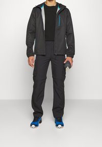 Veste softshell noire avec des fermetures éclair turquoise, associée à un pantalon cargo noir et des chaussures de sport bleues, tenant une bouteille d'eau grise.
