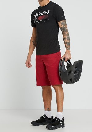 Mann trägt ein schwarzes Fox Racing T-Shirt und rote Shorts, hält in einer Hand einen schwarzen Fahrradhelm.