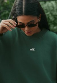 Groene trui met een ronde hals en geborduurd "self." logo. Model draagt schildpadkleurige zonnebril, met lang donker haar en lichte nagels.