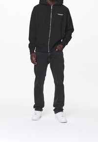 Felpa nera con zip e tasche frontali, abbinata a jeans grigio scuro e sneakers bianche. La felpa presenta un piccolo logo sul petto.