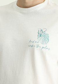 Biała bawełniana koszulka z grafika w kolorze teal przedstawiająca liść monstera oraz napisem "znajdź mnie pod palmami" w luźnej czcionce.
