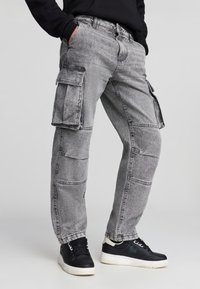 Pantaloni cargo grigi con molte tasche, una vestibilità ampia e dettagli di cucitura a contrasto. Indossati con sneakers nere. Materiale in denim leggero.