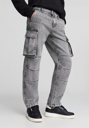 Pantaloni cargo grigi con molte tasche, una vestibilità ampia e dettagli di cucitura a contrasto. Indossati con sneakers nere. Materiale in denim leggero.