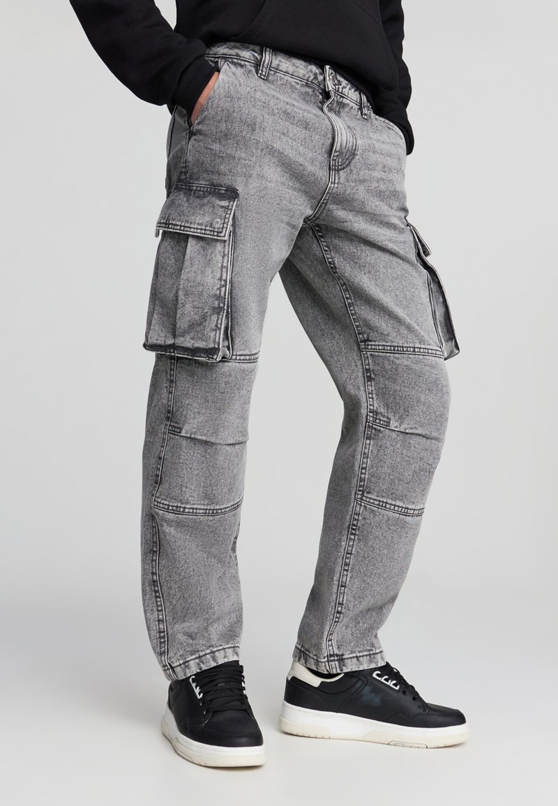 Pantaloni cargo grigi con molte tasche, una vestibilità ampia e dettagli di cucitura a contrasto. Indossati con sneakers nere. Materiale in denim leggero.