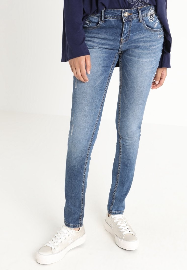 Jeans ajustados en denim azul claro, con un leve desgaste, detalles bordados y un corte slim, combinados con zapatillas blancas.