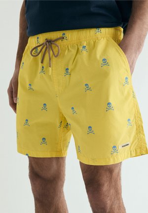 Hombre con pantalones cortos de baño amarillos con patrón de calaveras y huesos cruzados azules y cordón marrón, con la mano derecha en el bolsillo.