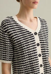 Cardigan a righe bianche e nere con scollatura a V profonda, maniche corte e cinque bottoni neri davanti, realizzato in tessuto a maglia testurizzato.