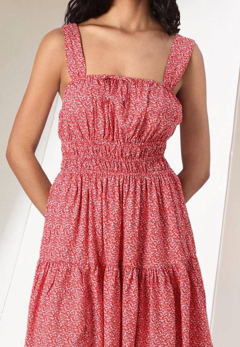Femme portant une robe rouge sans manches avec un imprimé floral blanc, un corsage élastique froncé et de larges bretelles.