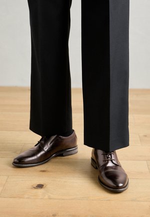 Braune Lederdressschuhe mit glatter Oberfläche, Schnürsenkeln und runder Zehenform. Kombiniert mit schwarzen Hosen, auf einem Holzboden.