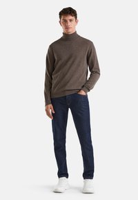 Maglione a collo alto marrone in maglia morbida, abbinato a jeans blu scuro. Il modello indossa sneakers bianche, con le mani nelle tasche, design semplice.
