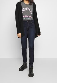 Czarny kardigan zapinany z przodu, noszony na szarej koszulce z grafiką przedstawiającą artwork zespołu, w połączeniu z ciemnymi rurkami i czarnymi botkami za kostkę.