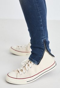 Canvas sneakers in off-white with red and blue accents, paired with dark denim jeans featuring zipper details at the ankles.