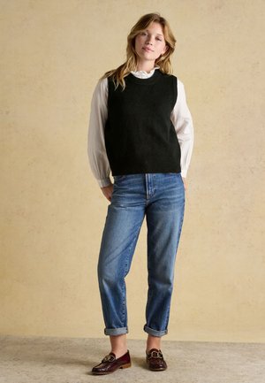 Ung kvinde iført sort ærmeløs sweater over en hvid bluse, blå bukser med optrukne ben og brune loafers, stående foran en beige baggrund.