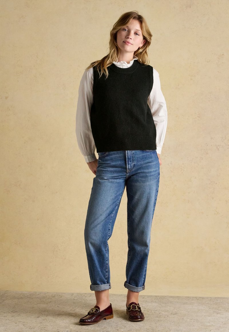 Junge Frau trägt schwarzen ärmellosen Pullover über weißer Bluse, blaue hochgekrempelte Jeans und braune Loafer, steht vor beigem Hintergrund.