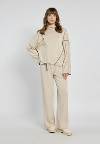 Beżowy oversize sweter z wysokim kołnierzem, ukośnymi szwami i zamkowymi akcentami, w połączeniu z pasującymi spodniami o szerokich nogawkach oraz butami na platformie.