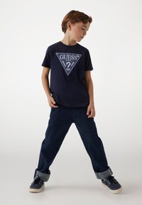 T-shirt cotone blu navy con logo triangolare in bianco e azzurro; abbinato a jeans denim scuri, risvoltati alle caviglie, e sneakers blu navy.