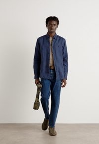 Blå knappjakke, beige skjorte, blå jeans og grå joggesko; bærer en grønn skulderveske. Enkel og uformell design.