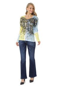 Langärmliges Hemd in Blau und Gelb mit grafischem Design, das eine Lilie zeigt. Kombiniert mit dunklen, ausgefransten Jeans und schwarzen High Heels.