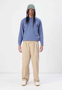 Chemise bleue à manches longues avec logo, pantalon beige ample et casquette verte. Des baskets blanches avec des rayures rouges complètent la tenue.