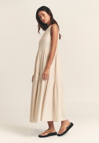 Béžové šaty bez rukávov s viacvrstvovým dizajnom, priedušnou látkou a jemnou textúrou. Kombinované s plochými čierno-hnedými žabkami.