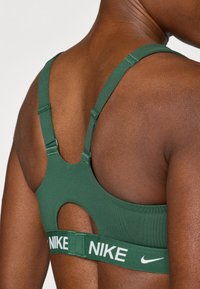 Πράσινο αθλητικό σουτιέν με ρυθμιζόμενους ιμάντες, σχέδιο με άνοιγμα στην πλάτη και λευκή λωρίδα με λογότυπο. Κατασκευασμένο από ελαστικό, απαλό ύφασμα.