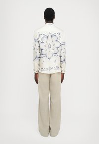 Weiße Bluse mit Blumenmuster und blauen Akzenten, Knopfverschluss, lange Ärmel; kombiniert mit beigen, weitgeschnittenen Hosen aus einem leichten Stoff.