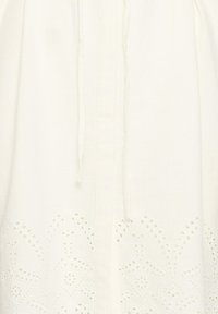 Tissu blanc avec couture verticale et motif floral découpé délicat brodé près de l'ourlet, présentant un détail de fine cordelette nouée.
