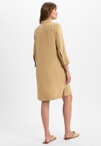 Beige linnen shirtjurk met driekwartmouwen, rugwerveldetail, zijsplitten en knoopmanchetten. Draagt met beige instapsandalen.