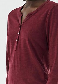 Maroon pitkähihaaninen henley-paita, joka on valmistettu pehmeästä materiaalista ja jossa on nappilista sekä pyöreä pääntie. Kankaassa on hienovarainen tekstuuri.