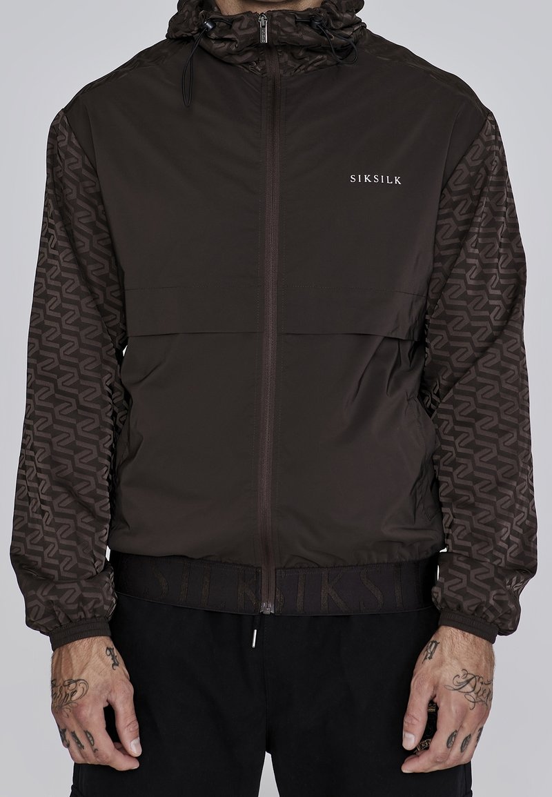 Veste à capuche marron foncé avec manches à motifs, fermeture éclair à l'avant et logo « SIKSILK » sur la poitrine, portée par une personne tatouée.