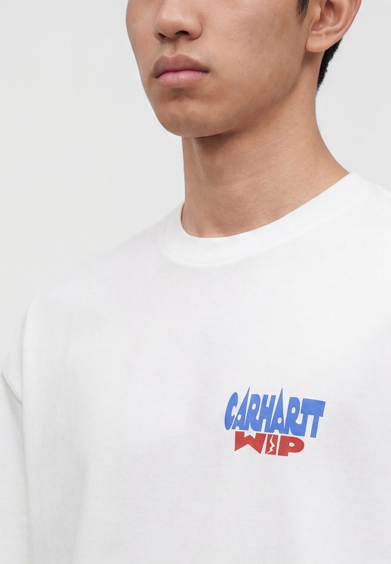 Uomo che indossa una maglietta bianca con il logo "Carhartt WIP" blu e rosso stampato sul petto a sinistra, mostrato dal collo al torace superiore.