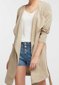 Cappotto lungo in lino beige con maniche lunghe, apertura frontale e design drappeggiato sopra una maglietta bianca e pantaloni shorts blu a vita alta con dettagli a bottoni.