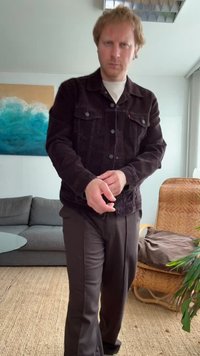 Veste en velours côtelé noir avec fermeture à boutons et deux poches poitrine, assortie à un pantalon sombre. La pièce comprend un canapé et une plante.