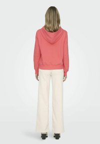 Sudadera color coral con cordón y bolsillo tipo canguro, combinada con pantalones de pierna ancha blancos. El conjunto presenta un ajuste relajado y una textura suave.