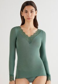 Long-sleeve top in forest green fabric, featuring a lace-trimmed V-neckline and lace accents at the cuffs, with a fitted silhouette.