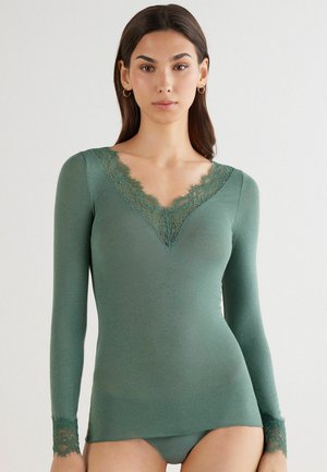 Langærmet top i skovgrøn stof, med en V-halsudskæring med blonder og blondeaccenter ved ærmerne, samt en tætsiddende silhuet.