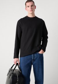 Camisa negra de manga larga con cuello redondo, combinada con jeans de mezclilla azul. El modelo sostiene un bolso de cuero negro.