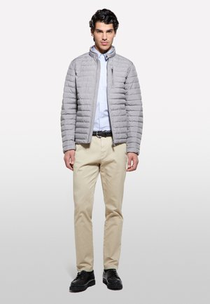 Giovane uomo in piedi che indossa una giacca trapuntata grigio chiaro, camicia bianca con bottoni, pantaloni beige, cintura nera e scarpe nere.