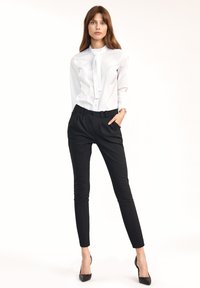 Biała koszula zapinana na guziki z kołnierzykiem „band”, w zestawieniu z eleganckimi czarnymi spodniami o dopasowanym kroju. Modelka ma na sobie czarne szpilki.