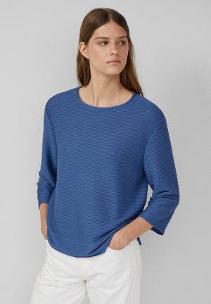 Blau gestreifter Langarmoberteil mit Rundhalsausschnitt. Der Stoff hat ein strukturiertes, geripptes Design. Das Model trägt weiße Hosen.
