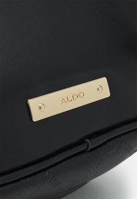 Černá texturovaná kabelka s pozlacenou kovovou destičkou s nápisem "ALDO", která zdůrazňuje jednoduchost designu a barevný kontrast.