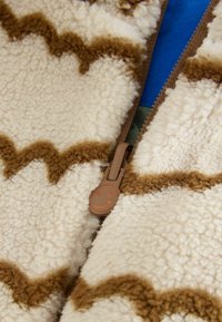 Bársonyos anyag, krémszínű alapszínnel és barna cikk-cakk mintával, barátságos mosolygós arccal ellátott cipzárral.