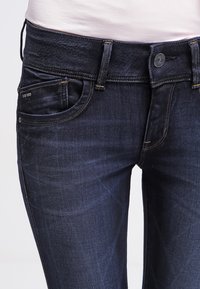 Vaqueros de mezclilla oscura con una textura suave, que presentan un diseño clásico de cinco bolsillos, costuras en contraste y un cierre frontal con botón.